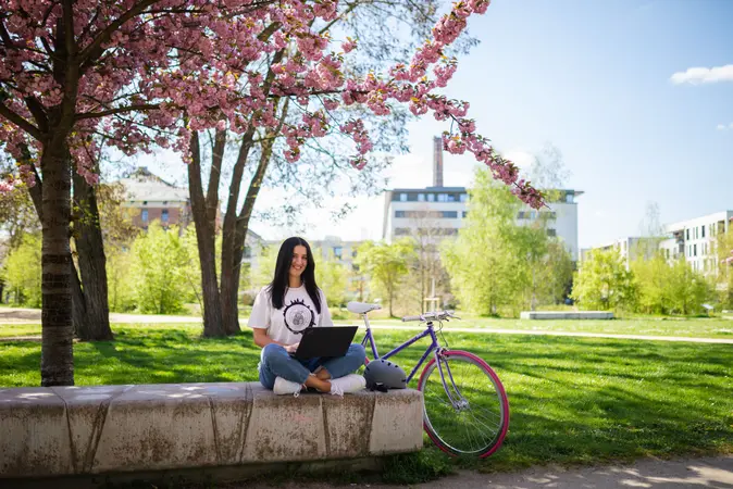 Studentin sitzt mit einem Laptop vor einem blhendem Baum im ERBA-Park vor einem Universit?tsgeb?ude