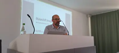 The photo shows Professor Jon Pineda, a man with short hair, a greying beard and glasses, standing behind a lectern and reading from his novel.