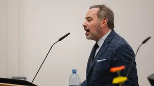 A photo of David Frum in profile. He stands behind a lectern and is talking into a microphone.